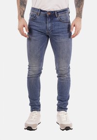 Uomo che indossa jeans blu slim-fit e sneakers bianche, in piedi con le mani rilassate lungo i fianchi, con avambracci tatuati visibili.