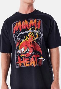 T-shirt nera in cotone con una grafica di un pesce rosso infuocato con grandi occhi, circondato da catene e le parole "MIAMI HEAT" in grassetto e colorato.