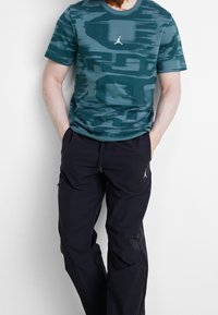 Mann in einem türkisfarbenen, gemusterten kurzärmligen Hemd und schwarzen Jogginghosen, mit den Händen in den Taschen, steht vor einem schlichten hellen Hintergrund.