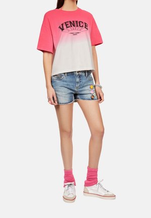 Różowy top crop z gradientem i grafiką "VENICE"; zestawiony z jasnymi szortami jeansowymi z kolorowymi ozdobnymi guzikami oraz białymi sneakersami.