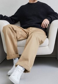 Przesadnie duży czarny sweter o fakturze, khaki luźne spodnie i białe sneakersy, siedzący na jasnym krześle na neutralnym tle.