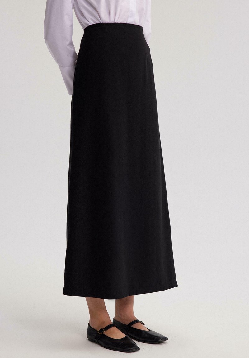 Femme portant une jupe longue noire taille haute associée à des chaussures Mary Jane noires et un chemisier blanc à manches longues rentré.