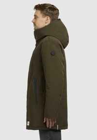 Brown hooded parka with a smooth texture, featuring zippered side pockets and a round logo patch on the shoulder. Casual, mid-length design.