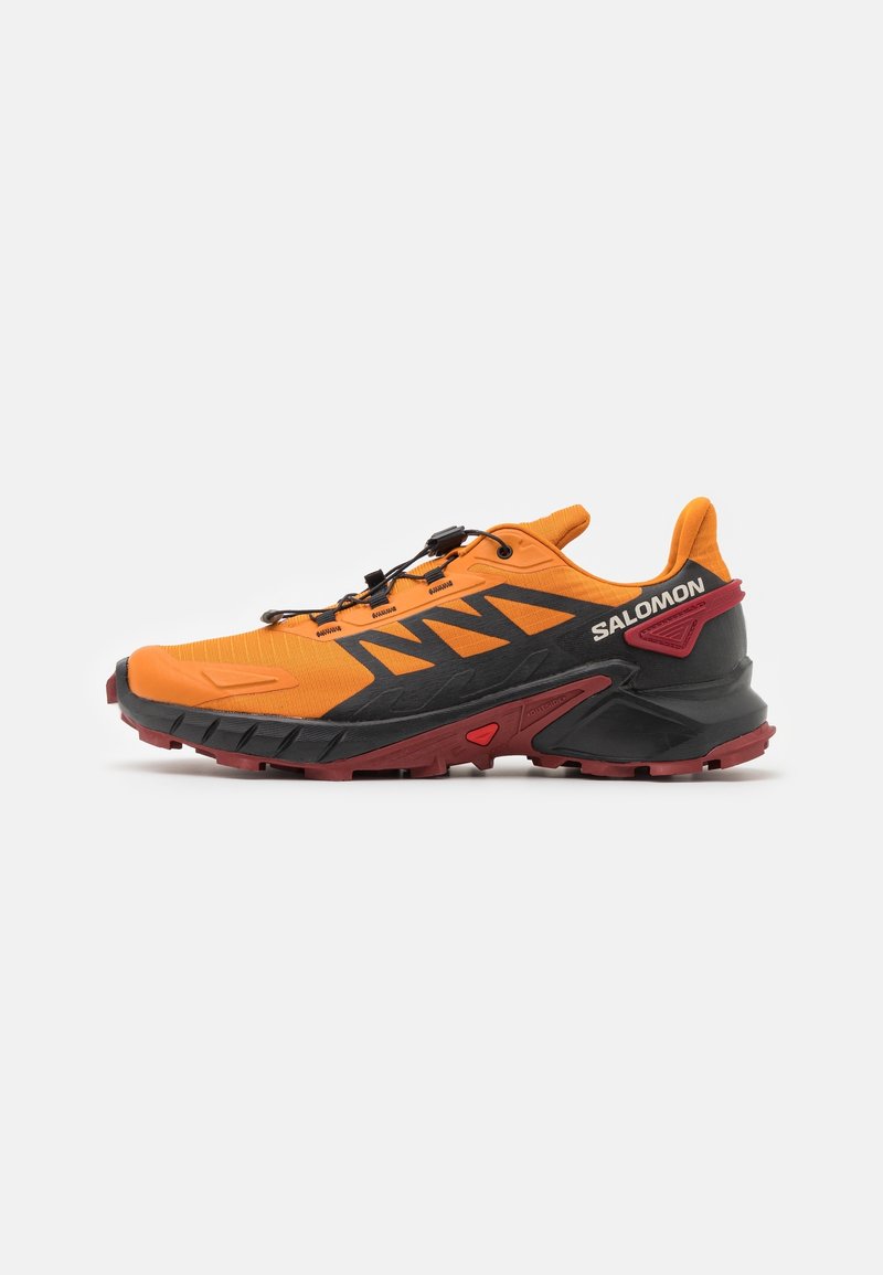 Chaussure de trail running Salomon en orange vif et noir avec semelle rouge, système de laçage rapide et semelle robuste pour une utilisation en extérieur, présentée de profil.