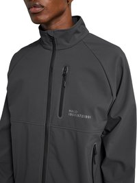 Man draagt een donkergrijs jack met rits, hoge kraag en ritsen op de borst en zijkanten, tekst "HALO 196819721981" op de borst.