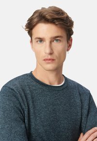Blauwe gebreide trui met textuur en ronde hals. Het model heeft kort golvend haar en een kleine hoopoorbel. De achtergrond is helder wit.