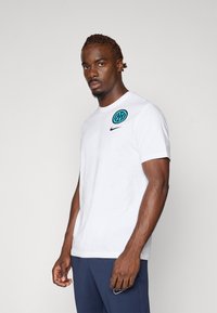 Hvid bomulds t-shirt med rund halsudskæring, forsynet med et sort Nike-logo og et blåt cirkulært design på venstre bryst. Glat tekstur.