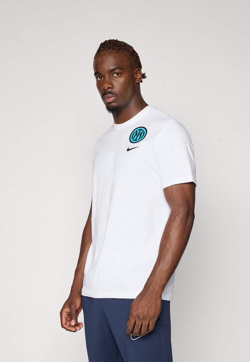 Hvid bomulds t-shirt med rund halsudskæring, forsynet med et sort Nike-logo og et blåt cirkulært design på venstre bryst. Glat tekstur.