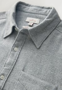 Pilkas flanelinis marškiniai su apykakle, sagų uždarymu ir krūtine kišene. Yra tekstūruota medžiaga ir prekės ženklo etiketės apykaklės viduje.