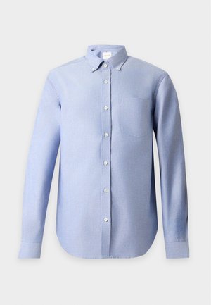 Homme debout portant une chemise bleu clair boutonnée, un pantalon marron foncé et des baskets en daim beige sur fond blanc.
