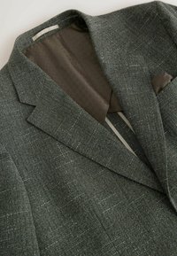 Veste texturée vert foncé avec un motif à carreaux subtil, doublure intérieure marron à motif et pochette marron assortie dans la poche poitrine.