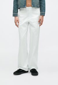 Άτομο που φοράει φαρδιά λευκή παντελόνα, μαύρα αθλητικά παπούτσια και ένα κοντό τζιν μπουφάν μπλε χρώματος που αφήνει ακάλυπτη τη μέση, στέκεται μπροστά σε ένα απλό φόντο.