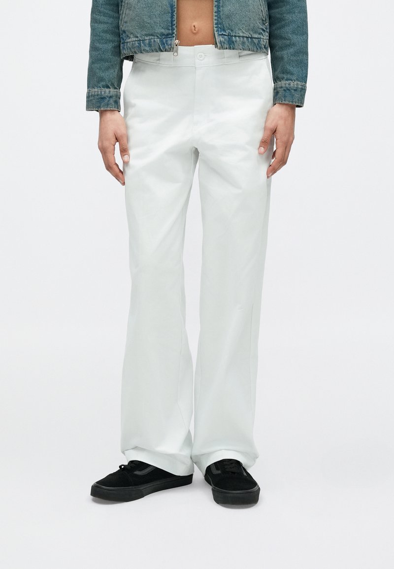 Persona che indossa pantaloni bianchi larghi, scarpe da ginnastica nere e una giacca di jeans blu corta con la pancia scoperta, in piedi contro uno sfondo semplice.