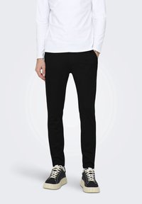 Pantalon noir slim en tissu texturé, associé à un haut blanc à manches longues et des baskets noires avec semelles et lacets blancs.