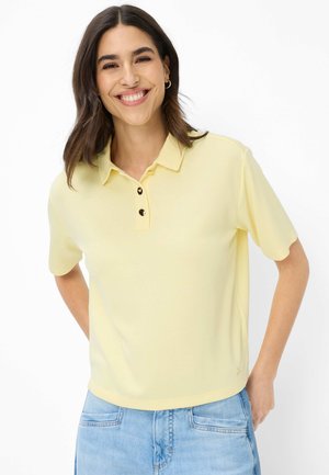 Donna sorridente con capelli scuri, che indossa una polo a maniche corte giallo chiaro e jeans azzurri, in piedi con le mani dietro la schiena.
