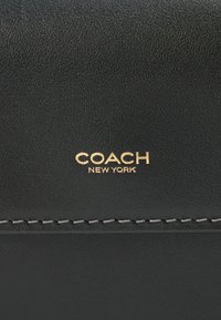 Črna usnjena denarnica z teksturirano površino in belim šivom. Ponaša se z zlatim logotipom "COACH NEW YORK" na vidnem mestu.