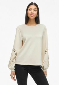 Felpa beige chiaro con scollo rotondo, maniche lunghe caratterizzate da motivi a foglia intagliati e tessuto morbido. Indossata con jeans neri.