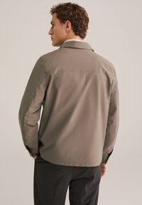 Homme de dos portant une veste collée marron clair et un pantalon gris foncé sur un fond beige uni.