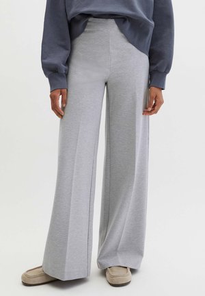 Person iført højtaljede, vidde bukser i lysegrå, beige slip-on sko og en mørk langærmet top med ærmer, der dækker håndleddene.