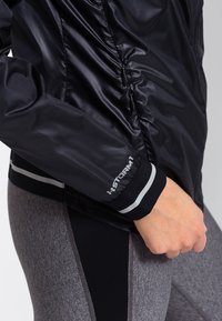 Czarna wodoodporna kurtka o eleganckim designie, z błyszczącym wykończeniem, dopasowanymi mankietami z szarym akcentem oraz logo "STORM 1" na rękawie.