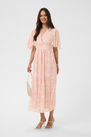 Femme portant une robe midi fleurie rose clair avec des manches volantées, tenant un petit sac à main beige et portant des sandales blanches à talons.