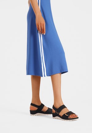 Donna che indossa un vestito blu con strisce laterali bianche e sandali zeppa neri con fascette alla caviglia, in piedi su uno sfondo bianco.