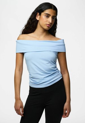 Kvinde med langt, mørkt, bølget hår iført en lyseblå off-shoulder-top og sorte bukser, stående mod en ensfarvet hvid baggrund.