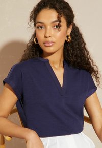 Navy-blaues kurzärmeliges Oberteil mit V-Ausschnitt. Weiches Material mit glatter Textur. Modell trägt einen hellen Rock und sitzt auf einem Stuhl.