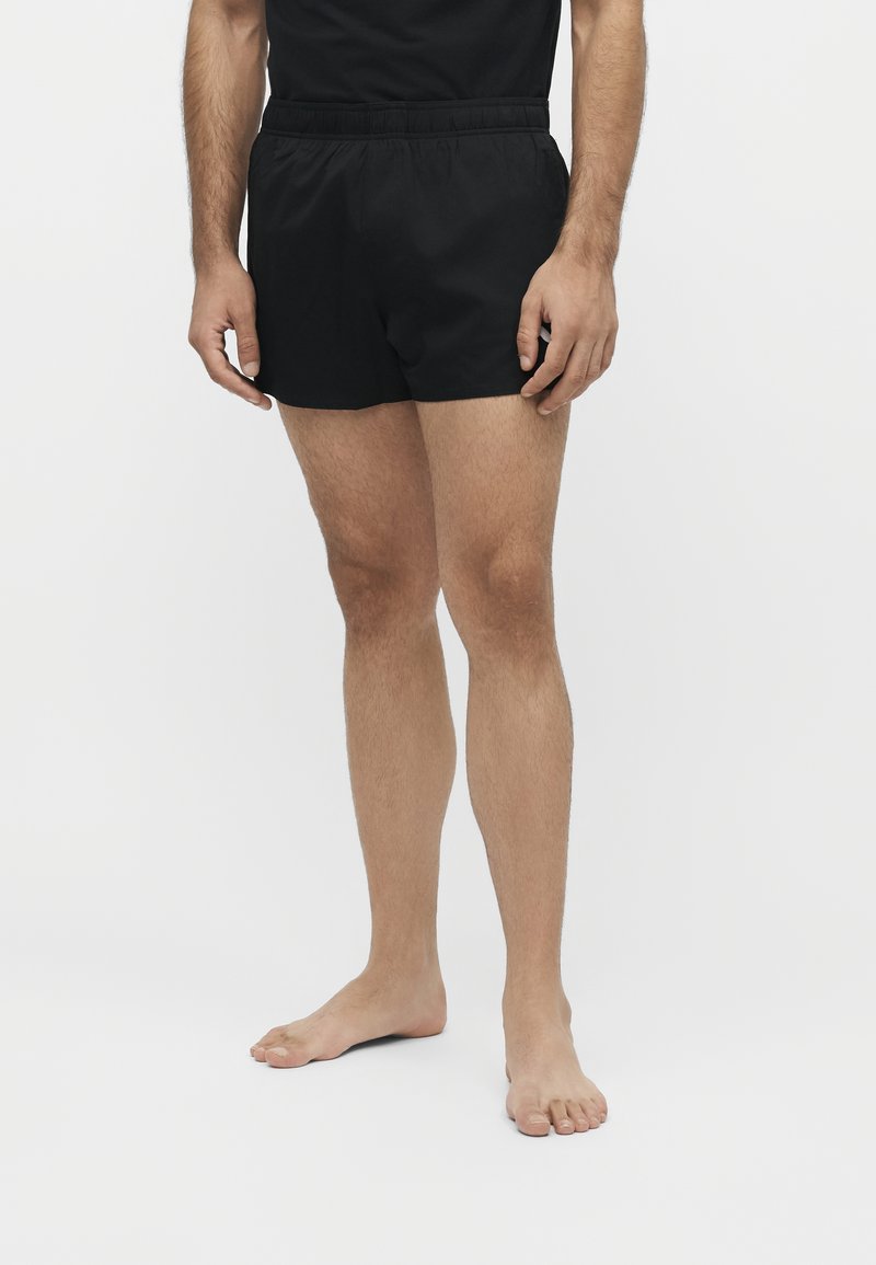 Uomo in piedi a piedi nudi, indossa pantaloncini sportivi neri e una maglietta nera su uno sfondo chiaro uniforme.