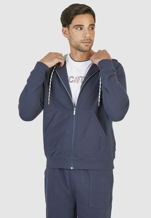 Haut à capuche zippé bleu marine en matériau polaire doux, avec capuche à cordon, poches avant et poignets côtelés. Présente un logo sur le bas à l'avant.