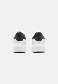 Baskets blanches avec semelles blanches épaisses et colliers de talon noirs vues de dos sur un fond clair uni.