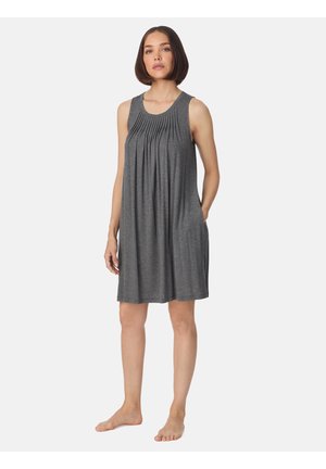 Vrouw staat blootsvoets en draagt een grijze mouwloze jurk tot op de knie met plooien aan de voorkant en zakken, tegen een effen lichte achtergrond.