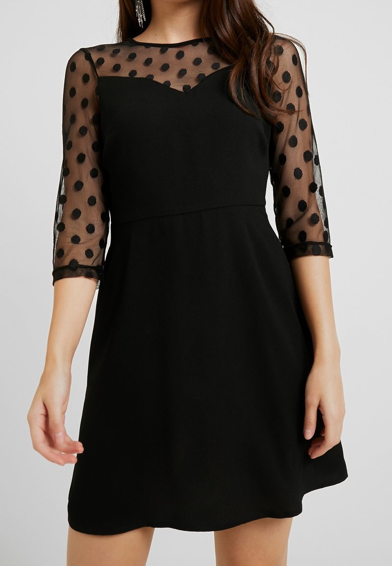 Mustat mekko, jossa on tiivis yläosa ja levenevä hame. Luihottavan läpinäkyvät, pilkulliset hihansuut lisäävät tekstuuria. Pyöreä pääntie luo naisellisen siluetin.