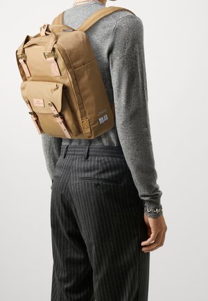 Persona con suéter gris y pantalones oscuros de rayas finas lleva una mochila beige con bolsillo frontal y correas ajustables.