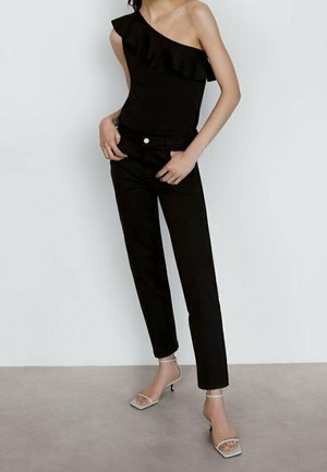 Femme portant un haut noir à une épaule avec volants, un pantalon noir à jambes droites, et des sandales à talons beige à lanières, debout contre un mur blanc.