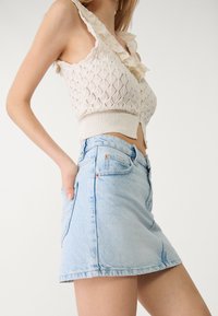 Gonna mini in denim azzurro chiaro con un design aderente, orlo sfrangiato e dettagli in rame, abbinata a un top in maglia bianca con accenti arricciati.