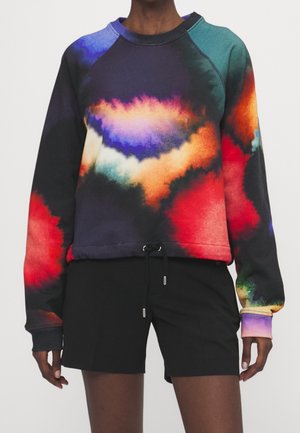 Osoba ubrana w wielokolorową bluzę tie-dye o odcieniach czerwieni, pomarańczy, fioletu i niebieskiego, w zestawieniu z czarnymi szortami.