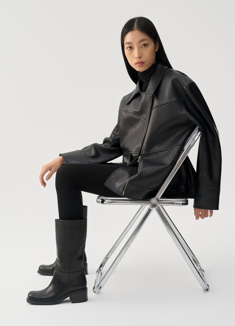 Giubbotto di pelle nera con zip e ampio colletto, abbinato a leggings neri a coste e stivali neri al ginocchio. Modella seduta su una sedia in cromo.