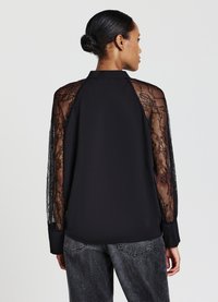 Blusa nera con sovrapposizione in pizzo sulle maniche, colletto e schiena in tessuto uniforme. Il materiale è leggero e ha una consistenza morbida.