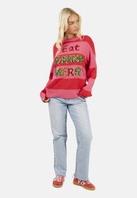 Maglione a righe rosa e rosso con lettere in paillettes "Eat DRINK MERR" in verde e rosso, abbinato a jeans azzurri chiari e sneaker rosse.