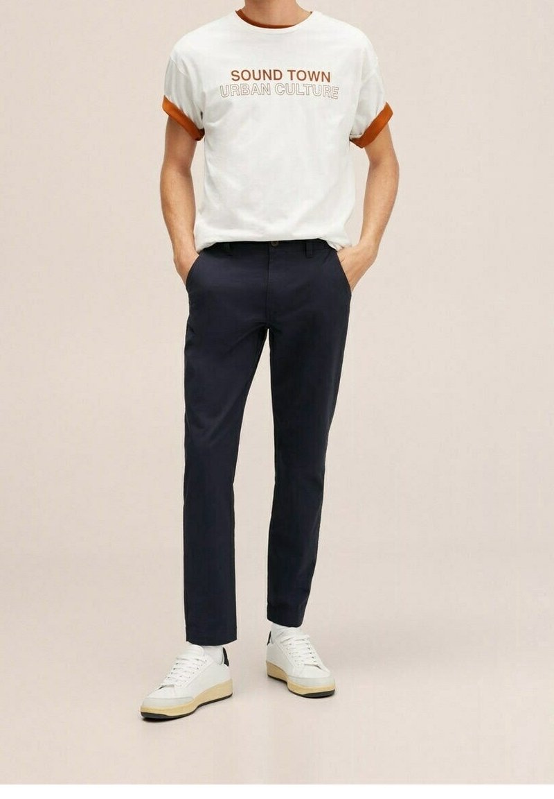 Homme portant un t-shirt blanc avec le texte "SOUND TOWN URBAN CULTURE", un pantalon bleu marine et des baskets blanches, debout les mains dans les poches.