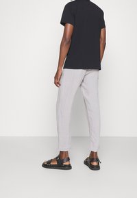 Osoba w jasnoszarych, lnianych spodniach, czarnej koszuli z krótkim rękawem i czarnych sandalach odkrytoe, stojąca na jednolitym, jasnym tle.