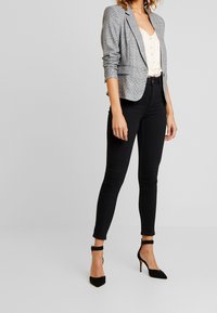 Grau karierter Blazer mit schmal geschnittenen schwarzen Jeans und schwarzen spitzen Absatzschuhen. Der Blazer hat einen Ein-Knopf-Verschluss und Dreiviertelärmel.