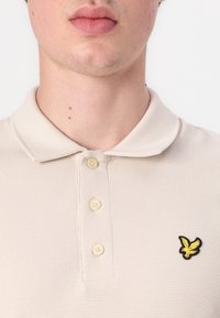 Beige polo tričko s texturovanou tkaninou, které má tříknoflíkovou légou a malé černobílé logo orla na hrudi.