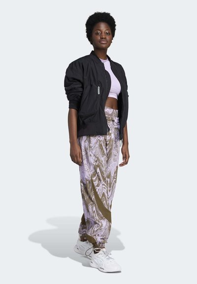 Fekete bomberdzseki, világos lila crop top, mintás levendula- és aranynadrág, fehér, fűzős edzőcipő, világos háttér előtt áll.