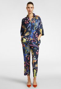 Camicia in satin blu navy con motivo floreale e pantaloni abbinati, caratterizzati da uccelli colorati e fogliame; completata da tacchi a punta arancioni.