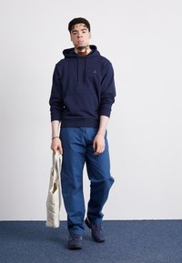 Granatowy hoodie, mieszanka bawełny, z kieszenią kangurzą. W połączeniu z niebieskimi spodniami i ciemnymi butami, trzyma beżową torbę na ramię.