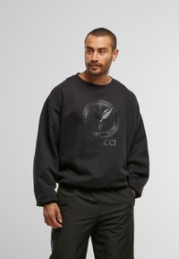 Felpa nera con un design centrale del logo metallico che presenta un motivo ad aquila. Il tessuto appare morbido, con una vestibilità comoda e polsini a costine.