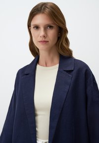 Blazer in lino navy con una vestibilità comoda, design aperto e colletto a bavero, indossato su una maglia leggera a coste. Tessuto liscio, dettagli minimi.