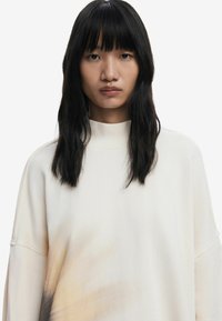 Felicità, il pullover oversized color crema presenta un colletto alto, maniche lunghe e una delicata trama sfumata che passa a un taupe chiaro nella parte inferiore.